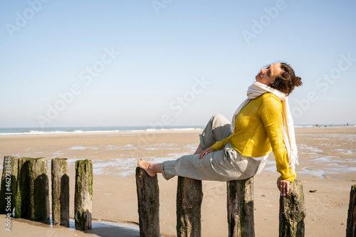 Mature woman with eyes closed sitting on wooden post at beach against clear sky photo