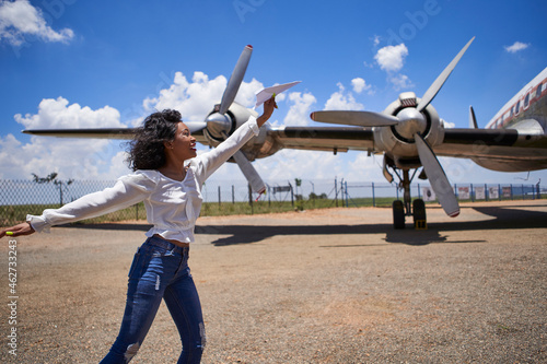 Cheerful young woman holding paper plane against propellers at airport on sunny day photo