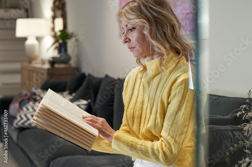 Mature woman in yellow sweater reading book on sofa at home photo