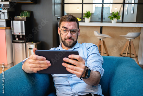 Businessman with raised eyebrow using digital tablet for video call while sitting on armchair at office cafeteria photo