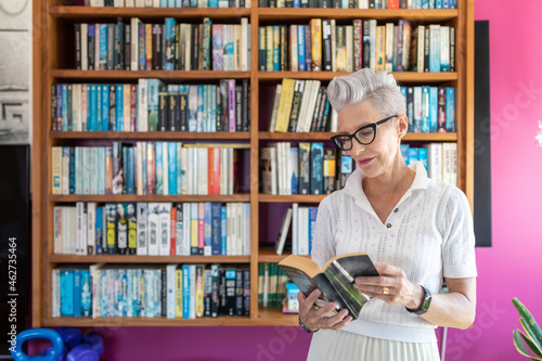 Active senior woman reading book while standing against bookshelf at home photo