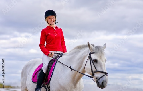 Portrait of a beautiful girl, young woman rider, equestrian on white horse in helmet and polo shirt, riding outdoors. Horseback riding sport. 