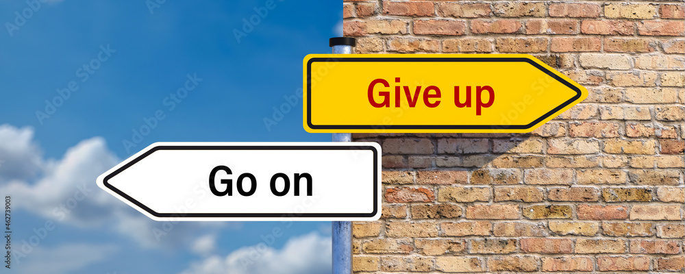 signpost pointing in two directions with the messages GIVE UP and GO ON in front of a brick wall and cloudscape signpost pointing in two directions with the messages GIVE UP and GO ON in front of a brick wall and cloudscape