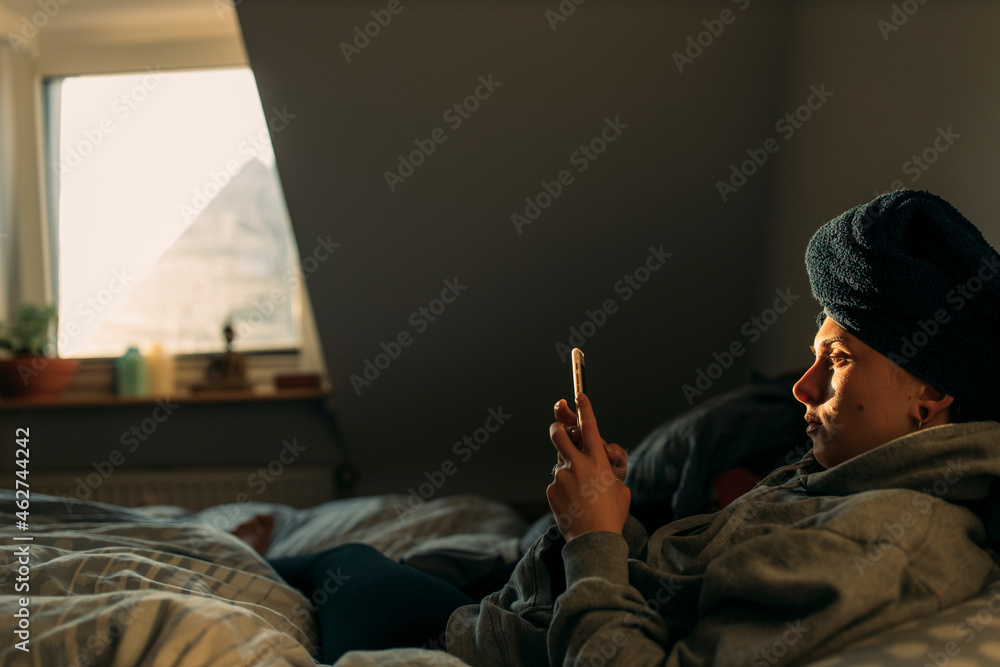 Young woman with head wrapped in a towel lying in bed at home using smartphone