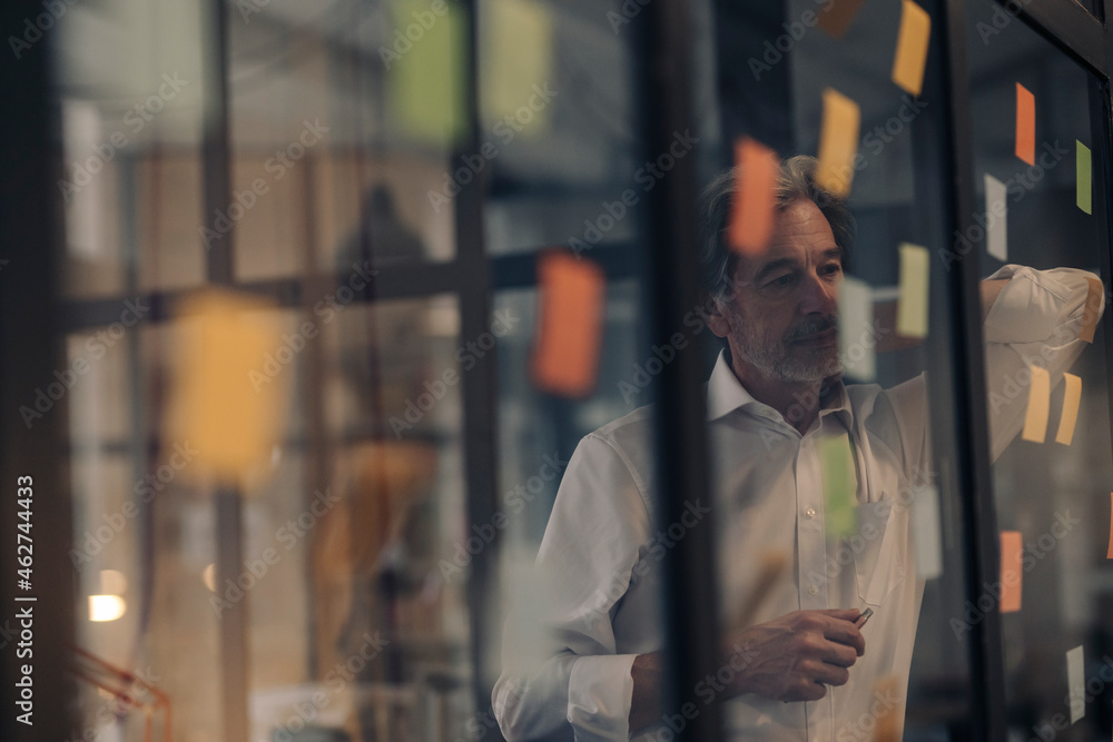 Senior businessman looking at adhesive notes at glass pane in office Senior businessman looking at adhesive notes at glass pane in office