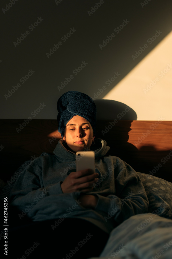 Young woman with head wrapped in a towel lying in bed at home using smartphone Young woman with head wrapped in a towel lying in bed at home using smartphone