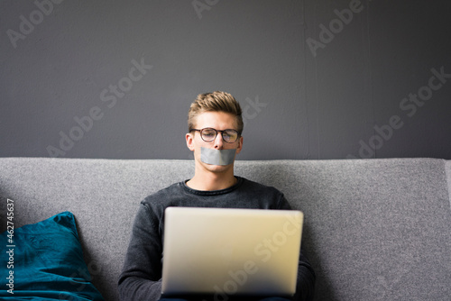 Young man with taped mouth sitting on couch using laptop