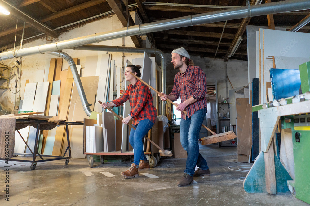 Playful craftswoman and craftsman riding on brooms in their workshop Playful craftswoman and craftsman riding on brooms in their workshop