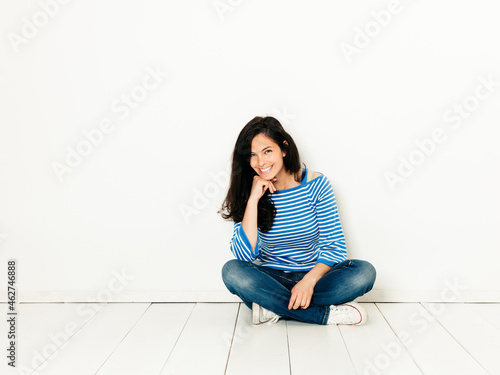 Beautiful young woman with black hair and blue white striped sweater sitting on the ground in front of white background