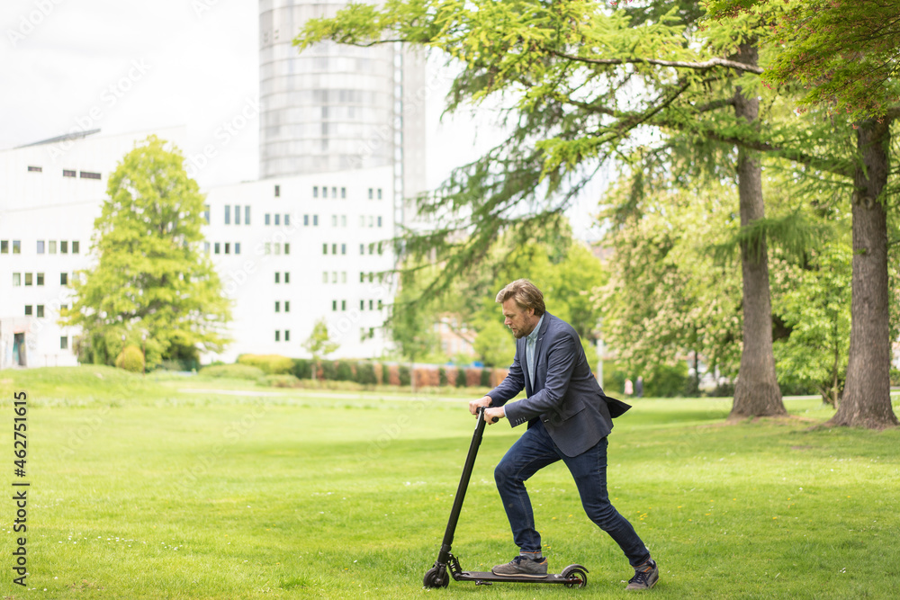 Businessman using E-Scooter on a meadow in city park, Essen, Germany Businessman using E-Scooter on a meadow in city park, Essen, Germany