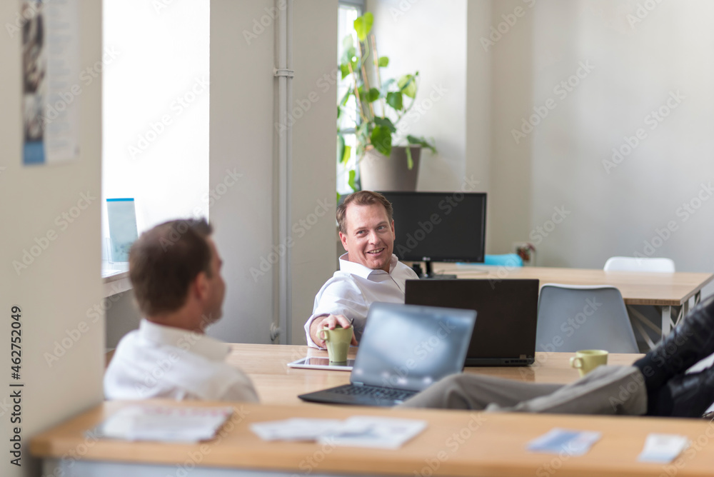 Two relaxed businessmen with laptops talking in office Two relaxed businessmen with laptops talking in office