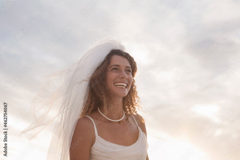 Cheerful young bride looking away while standing at beach against sky during sunset Cheerful young bride looking away while standing at beach against sky during sunset