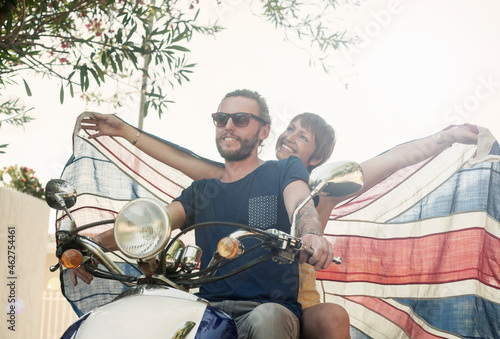 Smiling woman with arms outstretched holding national flag while sitting behind partner on motorcycle during summer photo