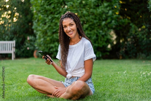 Smiling braided woman with mobile phone sitting cross legged in garden photo