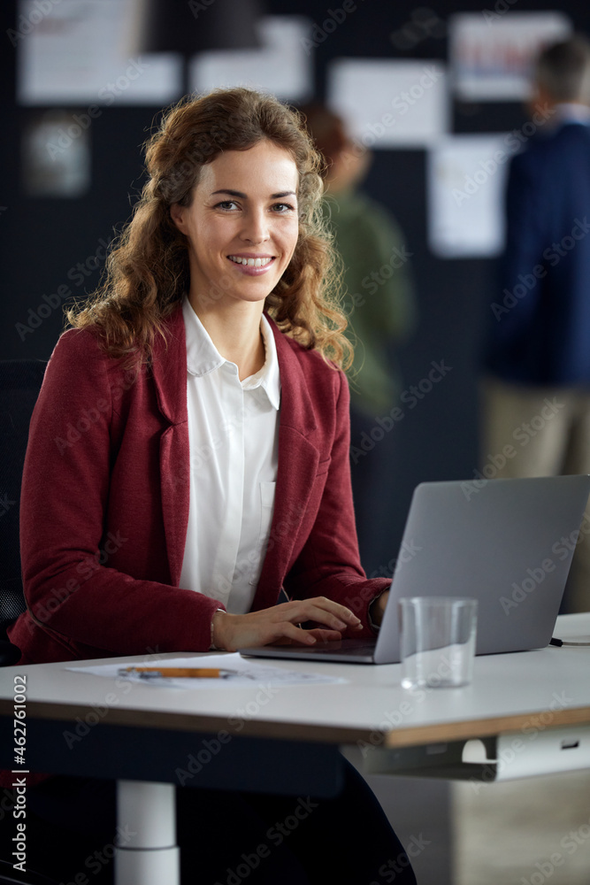 Portrait of smiling businesswoman using laptop at desk in office with colleagues in background Portrait of smiling businesswoman using laptop at desk in office with colleagues in background