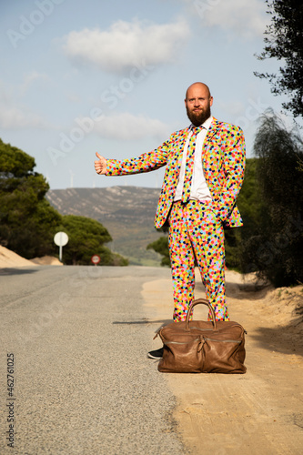 Hitchhiking man wearing suit with colourful polka-dots standing at roadside with travelling bag