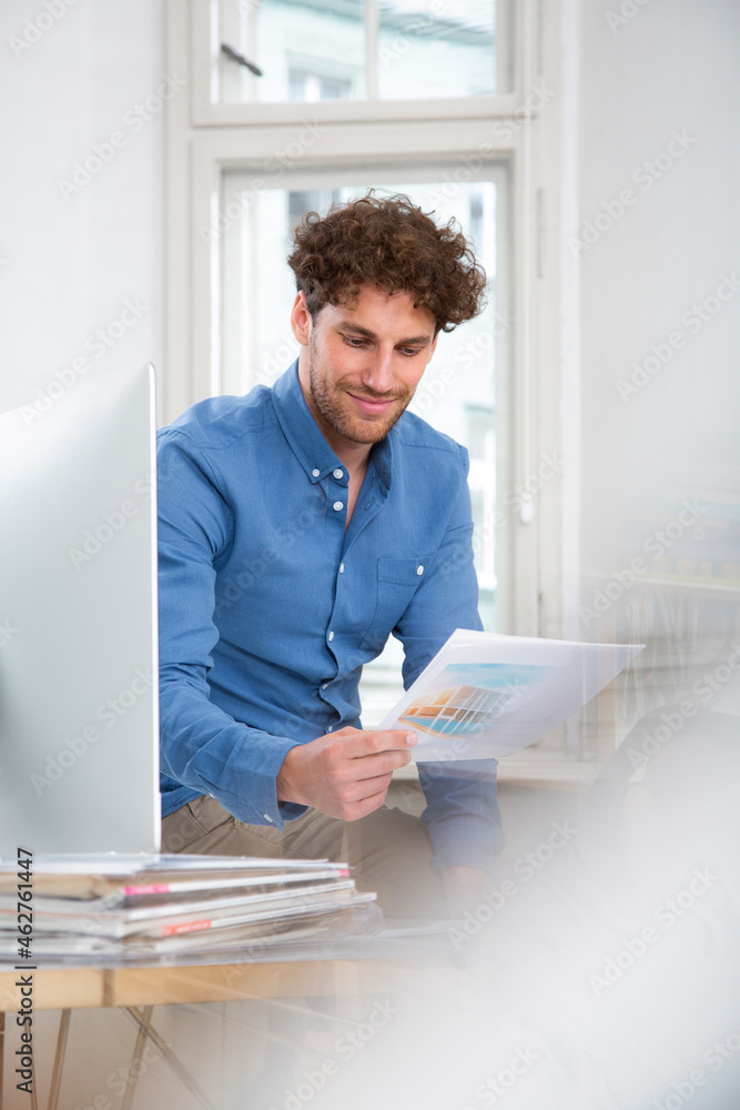 Male professional reading document sitting by computer in office Male professional reading document sitting by computer in office