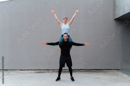 Woman with hand raised sitting on man shoulder standing with arms outstretched against wall photo