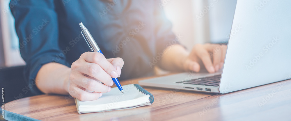 Business woman hand writing on a notepad with a pen and use a laptop computer in the office.