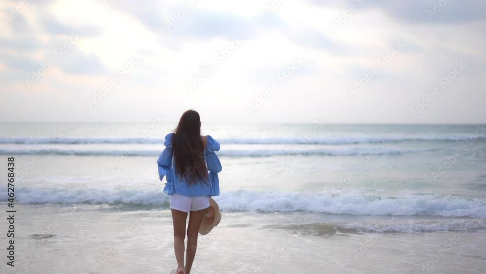 Asian Woman walking towards the sea on the beach turns around and raises arms up at sunset cloudy sky - vacation template back view Asian Woman walking towards the sea on the beach turns around and raises arms up at sunset cloudy sky - vacation template back view