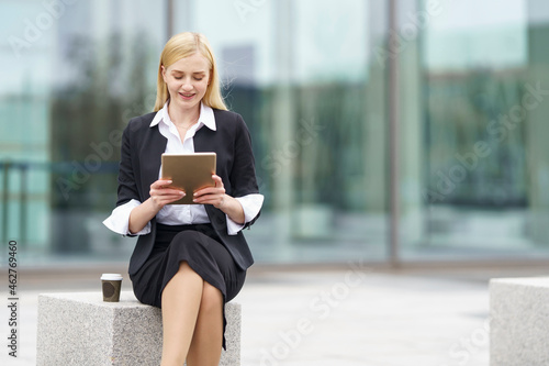 Blond businesswoman using digital tablet while sitting on bench against building photo