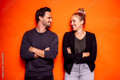 Happy mid adult couple looking at each other while standing with arms crossed against orange background photo