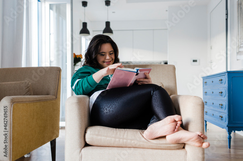 Happy young woman with down syndrome reading book while sitting on armchair at home photo