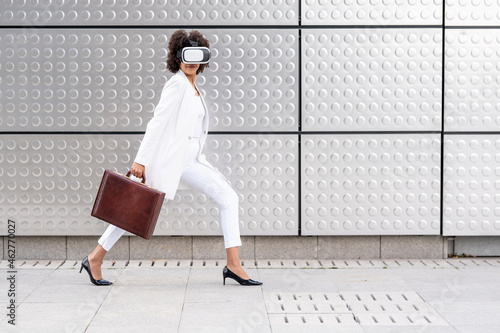 Young businesswoman with briefcase using virtual reality glasses while walking against silver wall photo
