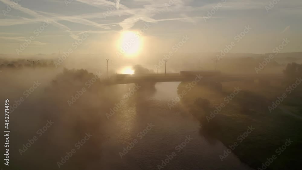Silhouette of tram crossing bridge over river, misty golden dawn sun rays, aerial shot Silhouette of tram crossing bridge over river, misty golden dawn sun rays, aerial shot