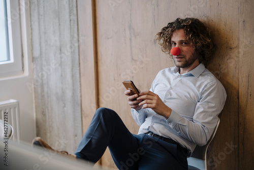 Portrait of businessman with red clown nose using cell phone in office photo