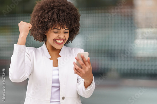 Cheerful businesswoman showing winning gesture while using mobile phone outdoors