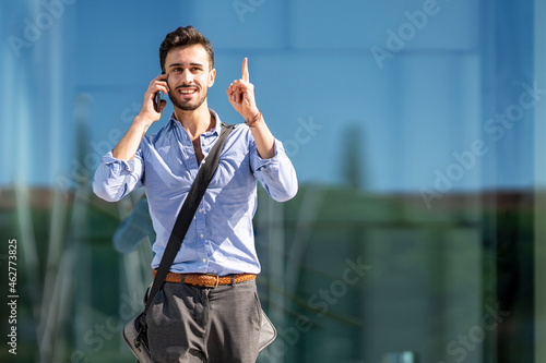 Businessman wearing crossbody bag pointing upward while talking on mobile phone outdoors photo