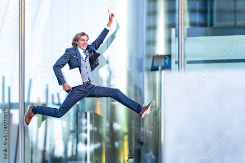 Excited male professional with laptop pointing while jumping against office building photo