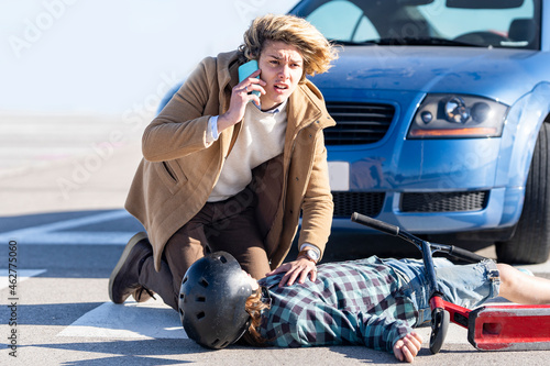 Young man calling help on mobile phone with boy in background lying on road after car accident