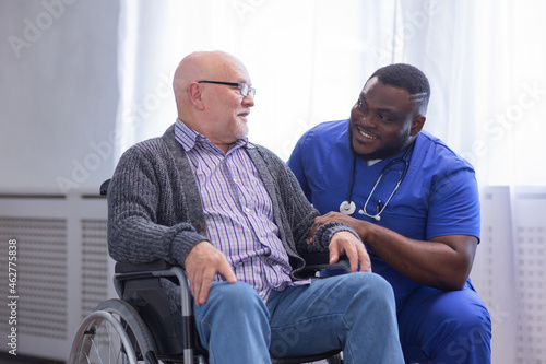 Fototapeta Naklejka Na Ścianę i Meble -  African-American caregiver and old disabled man in a wheelchair. Professional nurse and handicapped patient in a nursing home. Assistance, rehabilitation and health care.