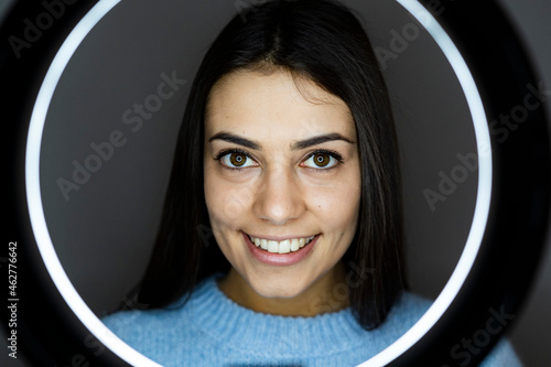 Young woman with illuminated circular camera flash smiling while standing against gray background photo