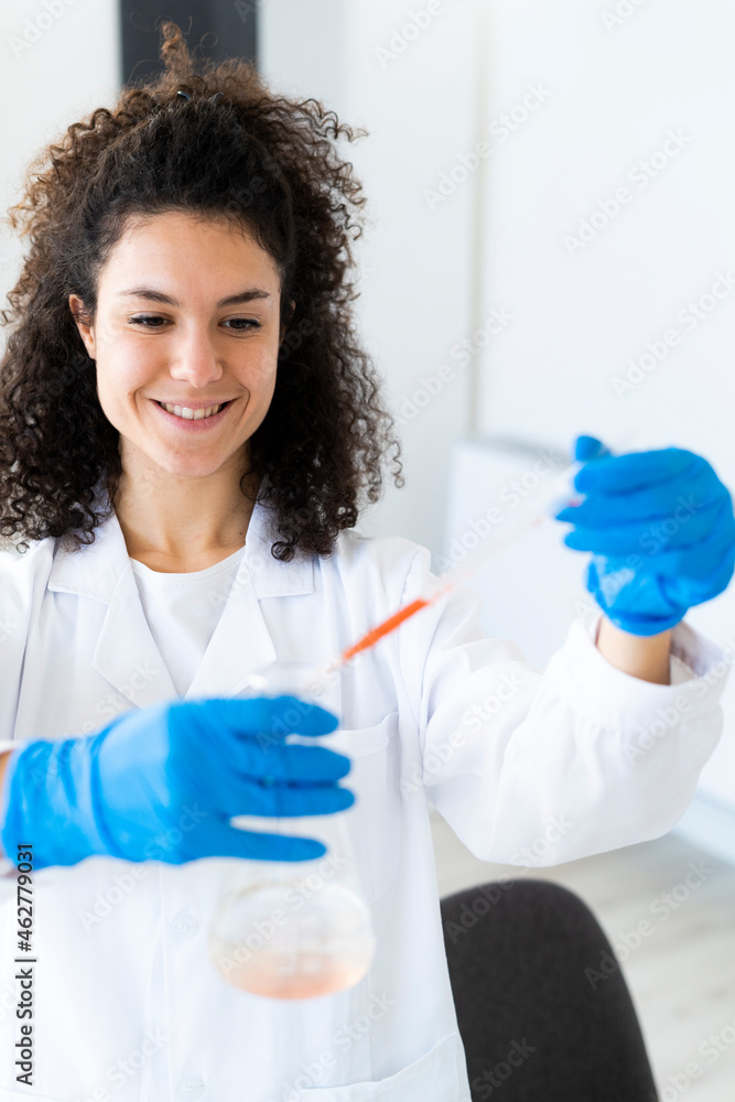 Smiling female doctor mixing solution in beaker through pipette at chemistry lab Smiling female doctor mixing solution in beaker through pipette at chemistry lab