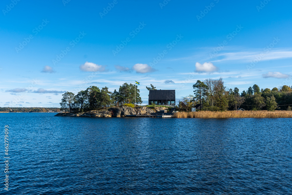 Swedish coast in autumn. Dark wooden small glass house with flag of Sweden. Beautiful colorful panoramic view of the rocky shores of the Baltic sea bay with thickets of reeds and evergreens pines.