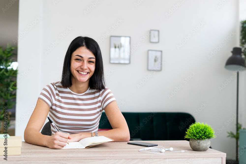 Cheerful woman with notebook and pen sitting at table in living room Cheerful woman with notebook and pen sitting at table in living room