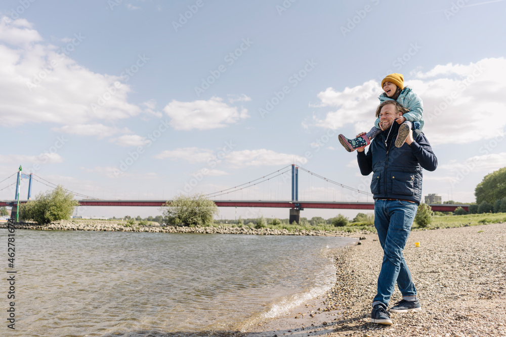 Father carrying daughter on shoulder while standing near river bank Father carrying daughter on shoulder while standing near river bank