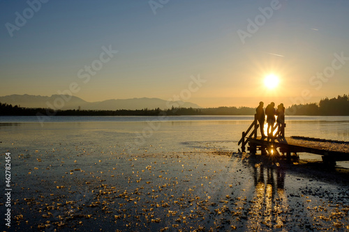 Silhouettes of three people standing on jetty at frozen Kirchsee by sunset, Bavaria, Germany photo