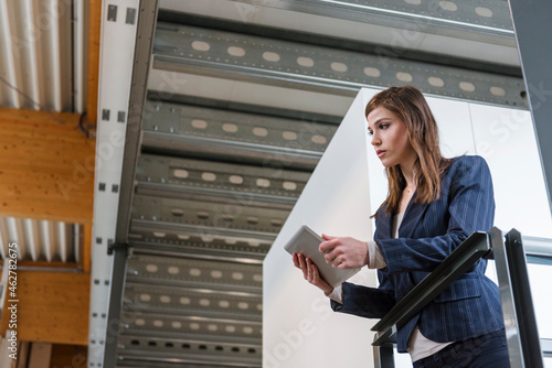Businesswoman holding digital tablet looking away while standing by railing in factory