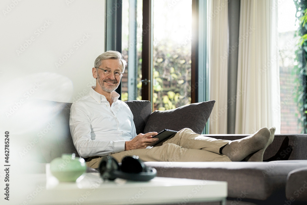 Smiling senior man with grey hair in modern design living room sitting on couch holding tablet Smiling senior man with grey hair in modern design living room sitting on couch holding tablet