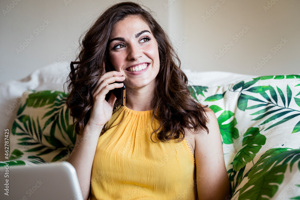 Smiling beautiful young woman talking on mobile phone while sitting with laptop and looking away at home Smiling beautiful young woman talking on mobile phone while sitting with laptop and looking away at home