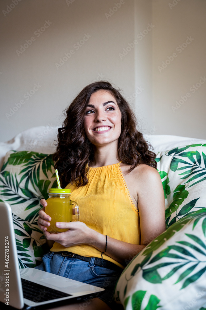 Thoughtful smiling woman holding juice while sitting with laptop on armchair at home Thoughtful smiling woman holding juice while sitting with laptop on armchair at home