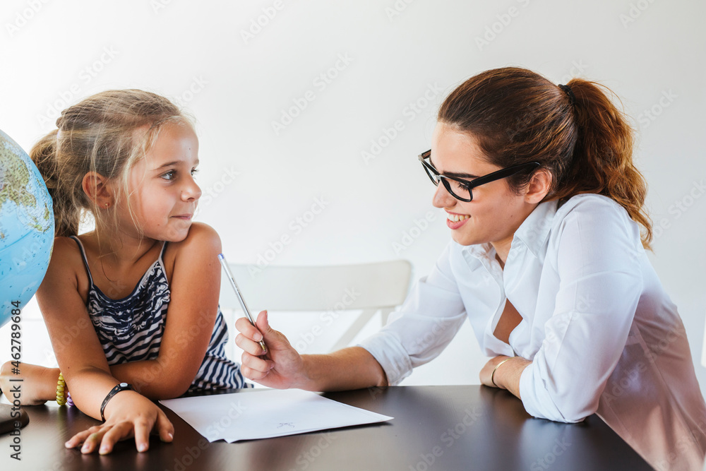Smiling teacher and schoolgirl sitting at desk with globe Smiling teacher and schoolgirl sitting at desk with globe