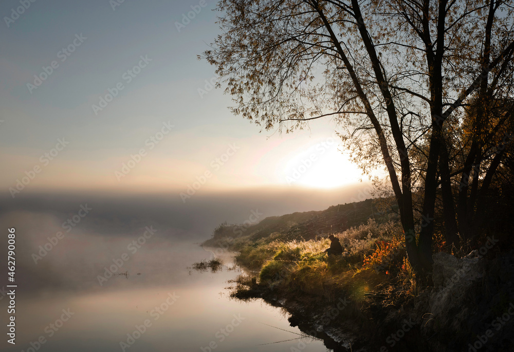 Fisherman with fishing rods on the bank of a quiet misty river in the early morning.