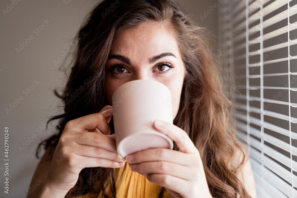 Close-up of young woman with long brown hair drinking coffee by blinds at home Close-up of young woman with long brown hair drinking coffee by blinds at home