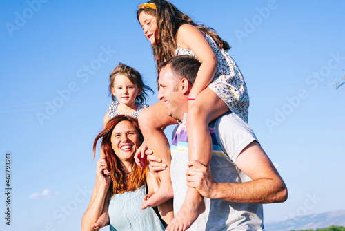 Cheerful parents carrying daughters on shoulders against clear sky during sunny day