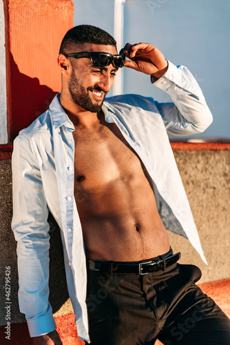 Smiling young man with shirt fully unbuttoned leaning on wall at rooftop photo